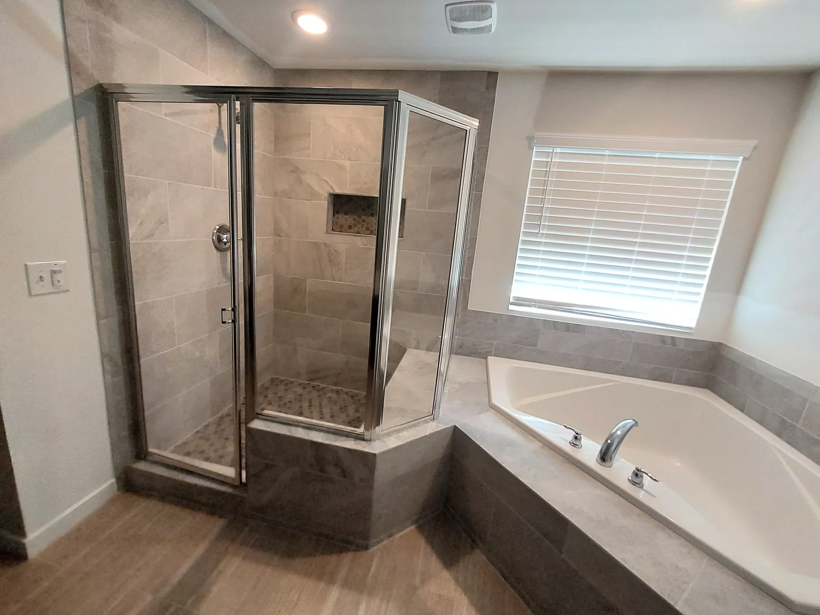 Glass-enclosed corner shower with pale stone patterned walls and a recessed niche beside a separate bathtub with a matching surround beneath a window.