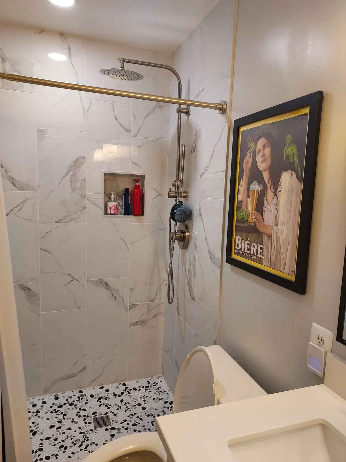 Shower with white stone patterned walls patterned with gray veining, an overhead showerhead and handheld sprayer, a recessed storage niche, recessed lighting, and a pebble patterned floor.