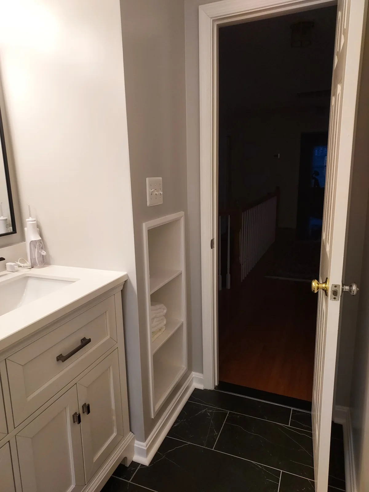 White recessed shelves holding folded towels beside a white vanity cabinet and bathroom doorway, with a dark stone patterned floor and white baseboards below.