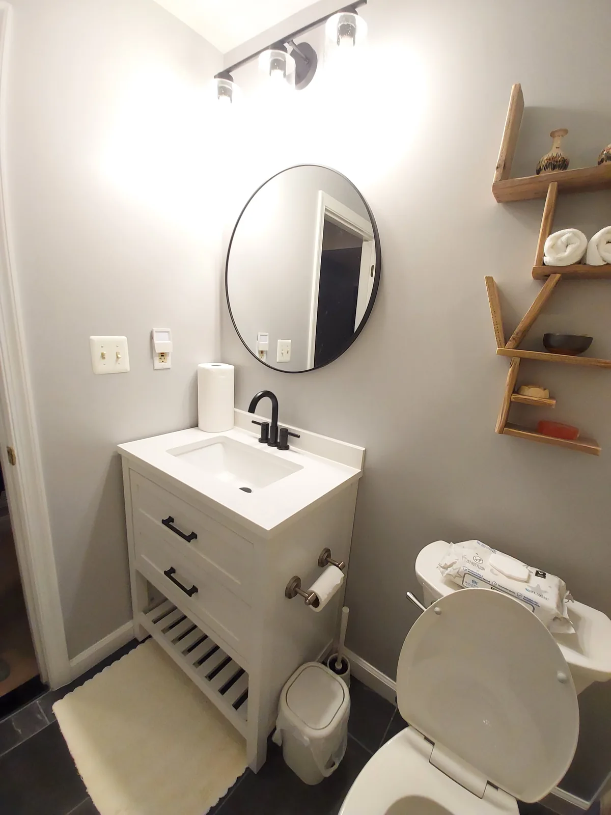 Compact white bathroom vanity with black faucet and drawer handles, an open lower shelf, a rounded mirror, a light fixture above, and staggered wood-tone wall shelves.