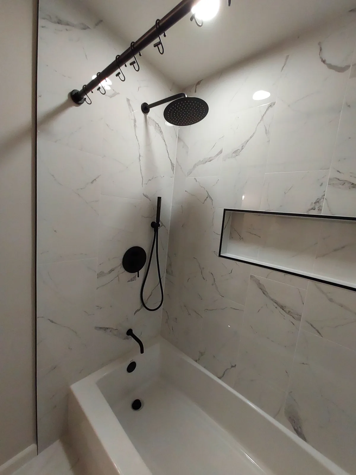 White bathtub with a stone patterned surround, a black overhead showerhead, a handheld sprayer, a black tub spout, and a wide recessed niche edged in black.