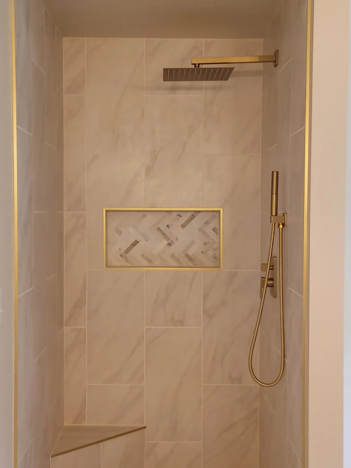 Shower with pale stone patterned walls, a square overhead showerhead, a handheld fixture, a gold-trimmed herringbone-pattern niche, and a corner seat.
