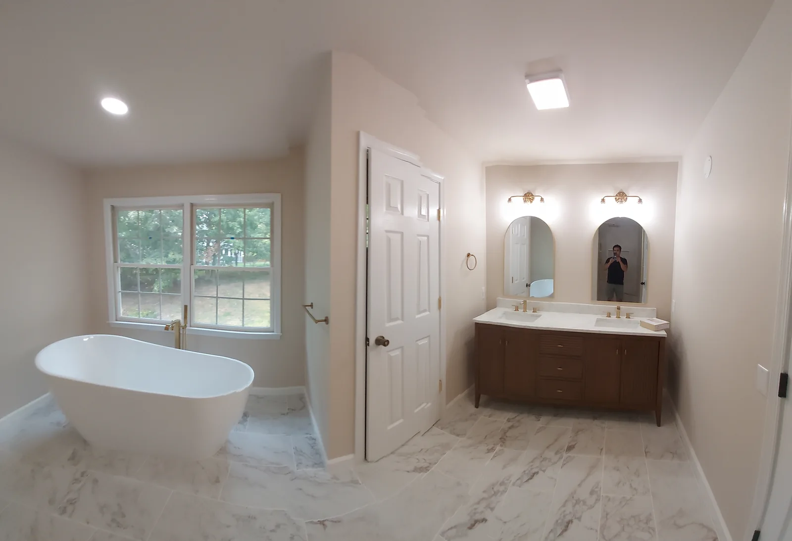 Bathroom with a freestanding white tub beneath two windows, a brown double vanity, arched mirrors, gold colored fixtures, and a pale floor with gray veining.